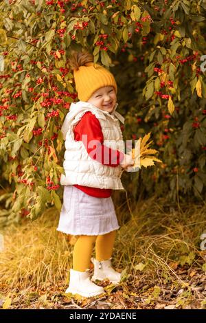 Un bambino allegro si erge tra un fogliame vivace, indossando un berretto giallo e un gilet accogliente. Sorride mentre tiene in mano foglie autunnali colorate, circondate da Foto Stock