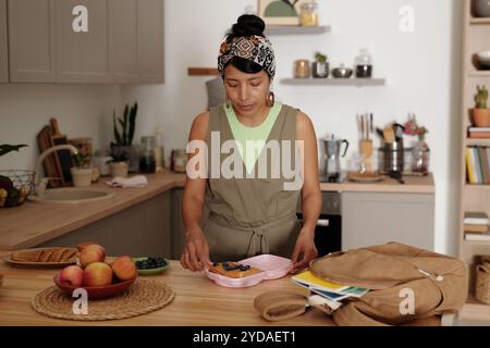 Giovane donna in una cucina rilassata che mette il cibo sul tavolo. Circondata da frutta fresca e utensili da cucina, mostra concentrazione e calma Foto Stock