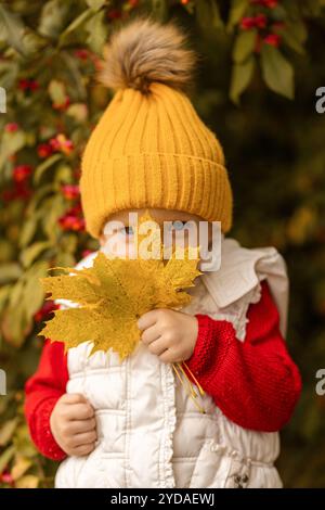 Un bambino allegro vestito con un maglione rosso e un accogliente cappello giallo gode della bellezza dell'autunno tenendo una grande foglia dorata in un giardino colorato. Foto Stock