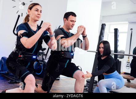 Giovani coppie sportive in tute ems sono impegnate in attività fisica in palestra con personal trainer. Foto Stock