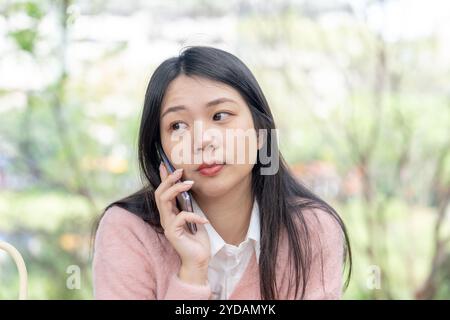 Una donna taiwanese dai capelli lunghi sui 20 anni che indossa un maglione rosa parla su uno smartphone mentre è seduta su una panchina del parco nel distretto di Xinyi, Taipei City, Ta Foto Stock