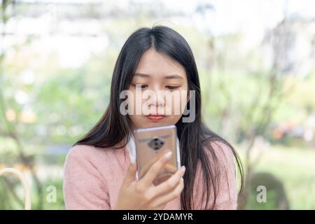 Una donna taiwanese dai capelli lunghi sui 20 anni che indossa un maglione rosa parla su uno smartphone mentre è seduta su una panchina del parco nel distretto di Xinyi, Taipei City, Ta Foto Stock