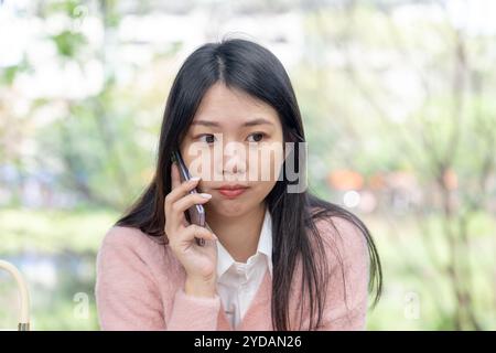 Una donna taiwanese dai capelli lunghi sui 20 anni che indossa un maglione rosa parla su uno smartphone mentre è seduta su una panchina del parco nel distretto di Xinyi, Taipei City, Ta Foto Stock