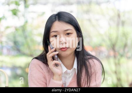 Una donna taiwanese dai capelli lunghi sui 20 anni che indossa un maglione rosa parla su uno smartphone mentre è seduta su una panchina del parco nel distretto di Xinyi, Taipei City, Ta Foto Stock