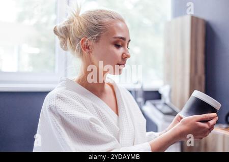 Una giovane donna attraente in un accappatoio guarda un barattolo di crema nella spa. Una ragazza affascinante tiene il mockup del barattolo in un beau Foto Stock