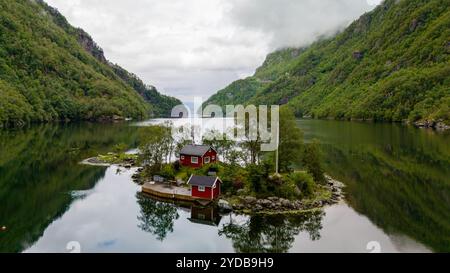 Una piccola isola annidata in un fiordo norvegese, con due cabine rosse con alberi verdi e montagne sullo sfondo. Lovrafjorden, Norvegia Foto Stock