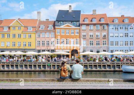 Una coppia siede su un muro di pietra che si affaccia su un trafficato canale di Copenaghen, ammirando la vibrante facciata di edifici storici e la vivace atmosfera di Foto Stock