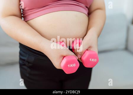 Donna in sovrappeso che si esercita per perdita di peso. Allenati con i manubri in posizioni strette a casa tua in salotto allegramente Foto Stock