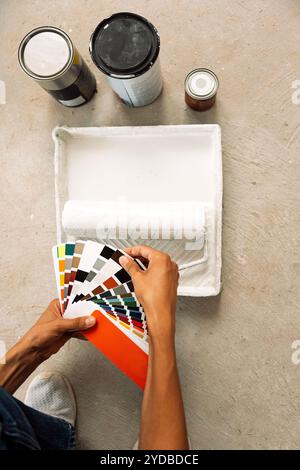 Primo piano di mani femminili che tengono una tavolozza di colori e lattine di vernice con il pennello a rullo. Una giovane donna seleziona il colore della vernice utilizzando Foto Stock
