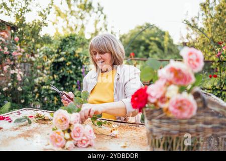 Una donna di mezza età è seduta a un tavolo e prende un mazzo di rose all'aperto. Una donna matura si prende cura dei fiori in t Foto Stock