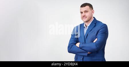 Giovane uomo d'affari di successo con le braccia incrociate che indossa una tuta blu con un sorriso delizioso e genuino che poggia su sfondo grigio Foto Stock