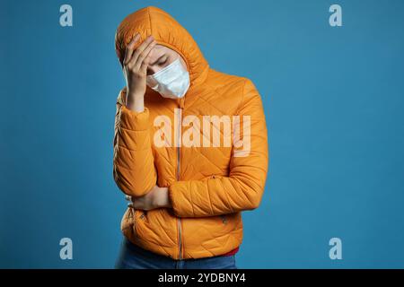 Donna che indossa cappuccio in una maschera protettiva e cappotto su sfondo blu. Modello ragazza in maschera medica e vestito di primavera, studio Foto Stock