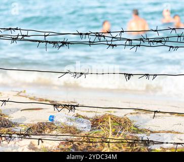 Vista attraverso il filo spinato su una spiaggia sporca piena di alghe, immondizia e nuoto persone Foto Stock