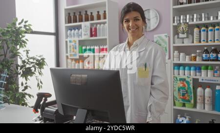 Sorridente, giovane, ispanica, farmacista donna si trova dietro il bancone in una farmacia ben organizzata, circondata da scaffali di vari prodotti e un com Foto Stock