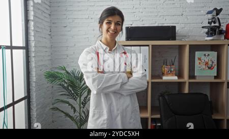 Una giovane dottoressa ispanica con i capelli bruna si trova in una clinica moderna con le braccia incrociate, mostrando un'accoglienza e una professio Foto Stock