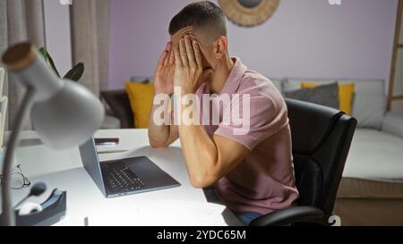 Un giovane in un ambiente domestico si siede a una scrivania con un notebook che appare stressato o frustrato, indicativo di uno scenario di lavoro da casa Foto Stock