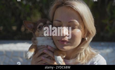 Una giovane donna bionda caucasica gode dell'affetto del suo animale domestico chihuahua in un parco urbano all'aperto, sottolineando uno stretto legame tra proprietario e Foto Stock
