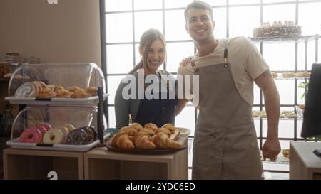 Fornai uomo e donna che lavorano insieme in una panetteria con pasticcini e ciambelle esposte al chiuso Foto Stock