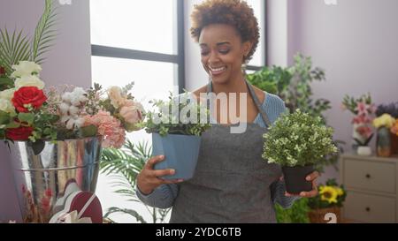 Fiorista sorridente afro-americana che tiene le piante all'interno di un negozio di fiori circondato da composizioni floreali assortite. Foto Stock