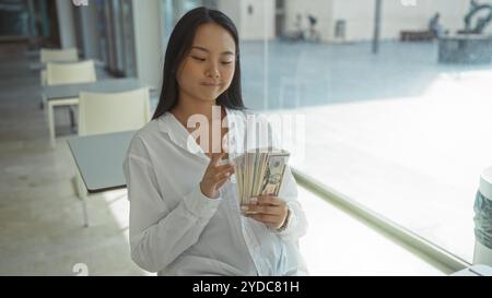 Giovane donna cinese che conta i soldi in un ufficio moderno, suggerendo successo finanziario e prosperità in un ambiente professionale. Foto Stock
