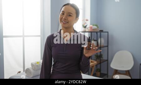 Una donna sorridente al chiuso tiene un prodotto cosmetico in un ambiente spa, mostrando bellezza e benessere in un ambiente di salone di ispirazione asiatica pieno di nat Foto Stock