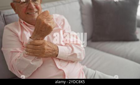 Un anziano uomo caucasico con occhiali e una camicia rosa si siede su un comodo divano in un luminoso soggiorno, sorridendo calorosamente con un comportamento rilassato. Foto Stock