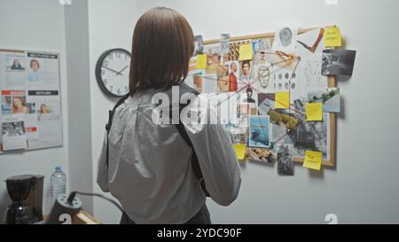 Una detective donna analizza le prove su un corkboard nella stanza delle indagini di un dipartimento di polizia. Foto Stock