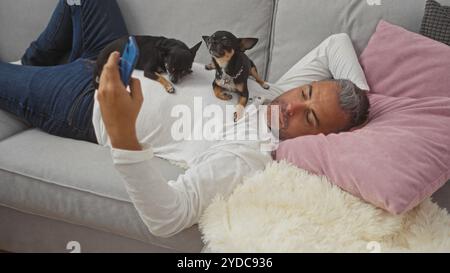 Uomo ispanico di mezza età che si rilassa in un soggiorno con due chihuahua appoggiati sul petto mentre utilizza uno smartphone. Foto Stock