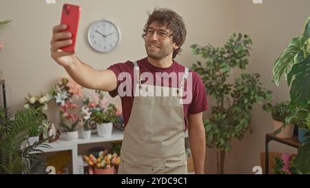 Un uomo ispanico sorridente con la barba scatta un selfie in un negozio di fiori al coperto, circondato da mazzi e piante colorate. Foto Stock