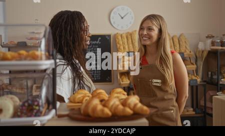 Donne che parlano all'interno della panetteria con pane e pasticcini in mostra circondate da scaffali e menu di lavagna sullo sfondo Foto Stock