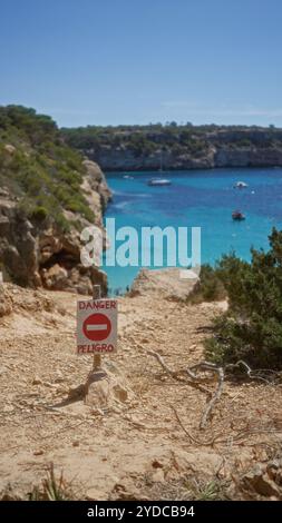 Cartello di pericolo che avvisa i turisti sulla scogliera rocciosa che si affaccia sulle acque turchesi di maiorca, spagna, sottolineando il paesaggio costiero panoramico ma pericoloso. Foto Stock