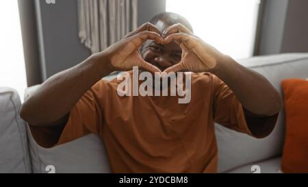 Un giovane uomo che forma il cuore con le mani seduto sul divano nell'accogliente soggiorno di un appartamento moderno. Foto Stock