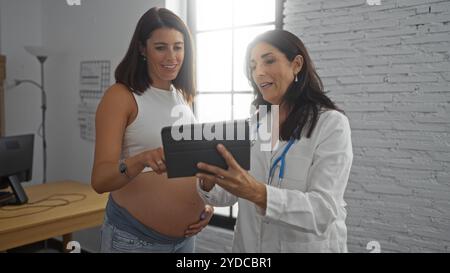 Donna incinta si consulta con un medico donna in una stanza clinica, rivedendo le informazioni mediche su un tablet insieme Foto Stock