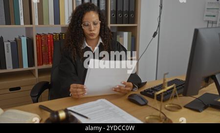 Una giovane donna giudice con i capelli ricci legge documenti alla scrivania del suo ufficio in un'aula di tribunale, circondata da libri legali e scale di giustizia Foto Stock