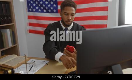 Giovane giudice che lavora in un ufficio con una bandiera americana sullo sfondo, usando un computer e controllando il suo telefono, circondato da libri legali e d Foto Stock