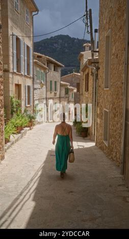 Donna che cammina lungo una stradina di valldemossa, all'aperto nella soleggiata maiorca, spagna, indossa un abito verde, porta un cesto intrecciato, con pietra Foto Stock