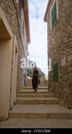 Donna che cammina attraverso una stretta strada di pietra a Port de valldemossa mallorca, spagna, tra edifici rustici sotto il cielo nuvoloso Foto Stock