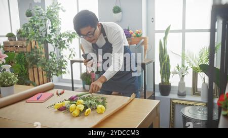 Un giovane asiatico organizza fiori in un luminoso negozio di fioristi mentre utilizza uno smartphone. Foto Stock
