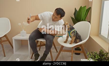 Un giovane asiatico fa un selfie con il suo cane in una sala d'attesa di una clinica veterinaria, seduto su sedie accanto a un tavolino con una pianta. Foto Stock