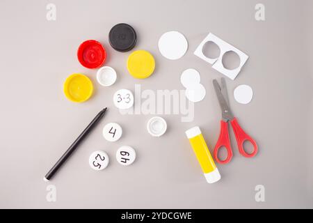 tappi per bottiglie in plastica con numeri scritti a mano e dati moltiplicati su sfondo grigio. Colla, forbici e pennarello, passaggio nel processo creativo fai da te, Foto Stock