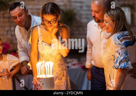 Una ragazza fa un desiderio e guarda la torta di compleanno. Una tenera adolescente con un abito elegante fa esplodere le candele per il suo compleanno. Genitori felici e rel Foto Stock