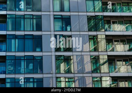 Un primo piano di un moderno edificio in vetro con finestre multiple e balconi. Le finestre riflettono il paesaggio urbano circostante, creando un motivo di blu A. Foto Stock