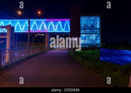 Un ponte pedonale illuminato con vivaci luci blu e viola si collega a un moderno edificio di vetro di notte. La scena è tranquilla e urbana, wi Foto Stock