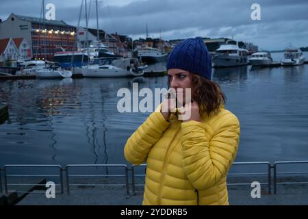 Donna di 30 anni che indossa una giacca imbottita gialla e un cappello di lana blu che tiene il colletto della sua giacca guardando lateralmente, con un porticciolo con le barche i. Foto Stock