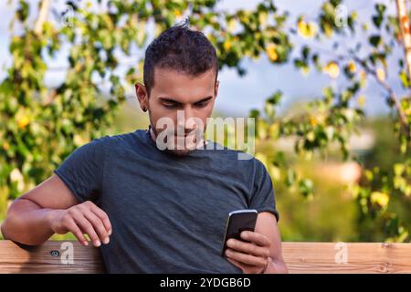 Il giovane è seduto su una panchina di legno in un parco, utilizzando il suo smartphone con un'espressione concentrata, godendosi l'ambiente tranquillo e restando conne Foto Stock