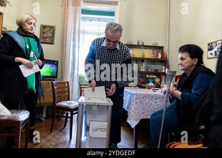 Tbilisi, Georgia. 26 ottobre 2024. Yuri colloca con attenzione il suo voto completato nella casella dei seggi elettorali, tenuta da un funzionario elettorale. Nani osserva silenziosamente, le sue mani appoggiate sulla sua stampella, mentre completano questo importante rituale di voto che rappresenta la loro speranza per il futuro pacifico e prospero della Georgia. (VX Photo/ Vudi Xhymshiti) crediti: VX Pictures/Alamy Live News Foto Stock