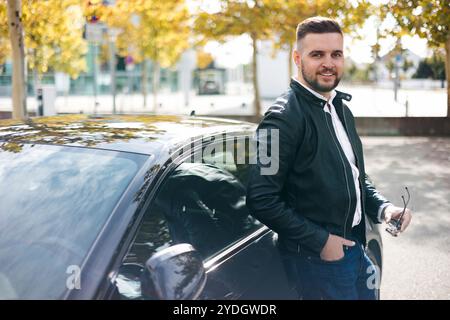 Un giovane uomo d'affari sta accanto alla sua auto di lusso di fronte all'ufficio. Elegante giacca in pelle nera da uomo con barba Foto Stock