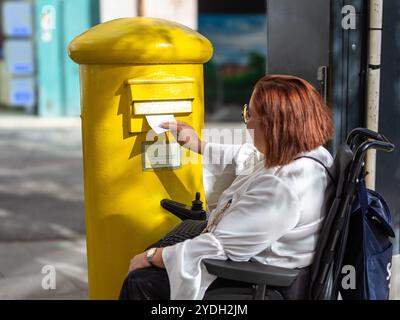 Donna che usa la sedia a rotelle elettrica per inviare una lettera nella cassetta postale pubblica Foto Stock