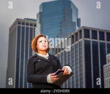 Una donna sta in piedi con sicurezza davanti a grattacieli torreggianti, tenendo un tablet in mano. Il cielo coperto e l'architettura moderna creano un vero e proprio mestiere Foto Stock