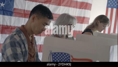 Concentratevi sul voto di una donna anziana con un uomo asiatico e una giovane donna caucasica, tutti votanti agli stand con la bandiera americana sul muro dietro Foto Stock